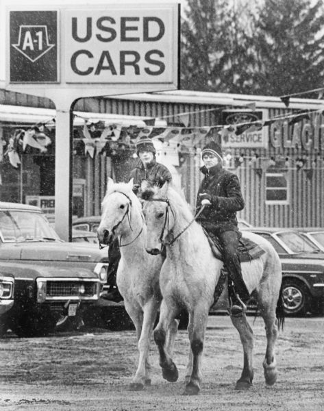 Two boys wearing jackets and knit hats are riding light-colored horses in town. Behind them is a sign overhead rows of automobiles that reads:  "Used Cars."
