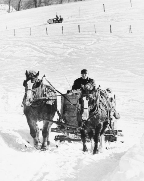 View down hill towards a man driving a large sled pulled by two draft horses up the hill through the snow. Further down the hill in the background is a snowmobile with two people.