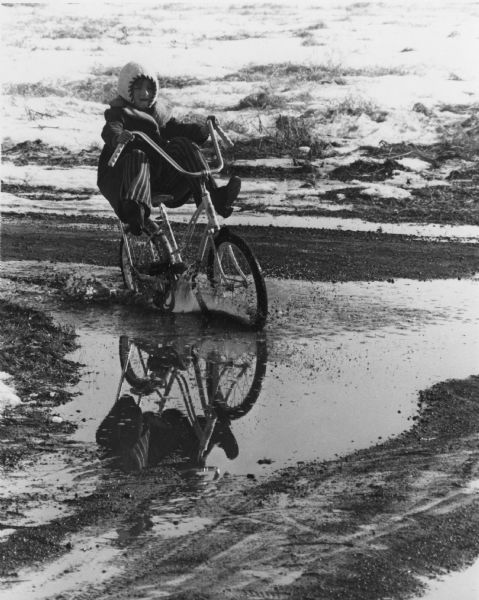 A girl is riding her bike through a puddle created by melting snow. She is holding her feet up off the pedals, away from the splashing water.