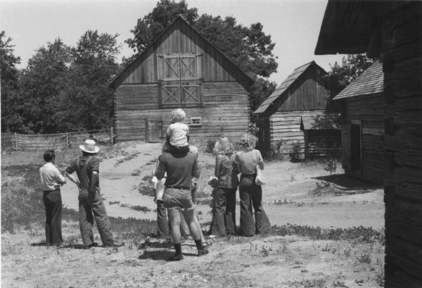 Rear view of a group of tourists at Old World Wisconsin viewing a barn and other farm buildings.