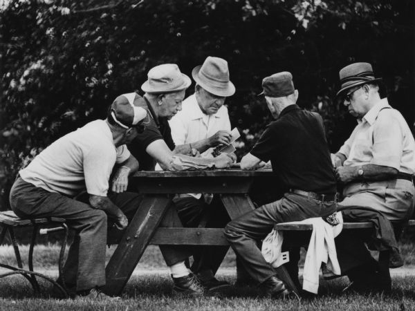 A group of senior citizens, all men, playing cards at a picnic table in an outdoor setting.