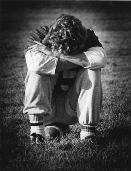 A young man is sitting on a football in the grass with his head down resting on his arms which are crossed over his knees.
