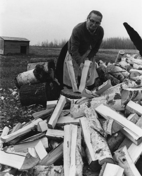 An older woman is splitting wood on a farm. She is wearing a dress and apron.