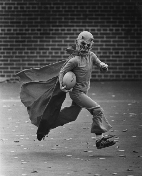 A boy dressed as a demon with a cape and holding a football is running in front of a brick wall.
