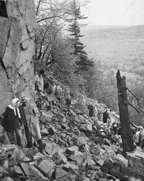 University students on a field trip to Devil's Lake State Park climb the trail up through the piles of talus (broken rocks) on the slope. A couple in the foreground is standing in front of a large outcropping. A forest is in the background.