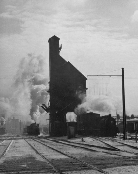 View down railroad tracks towards a tall building. Smoke is billowing from the locomotive.
