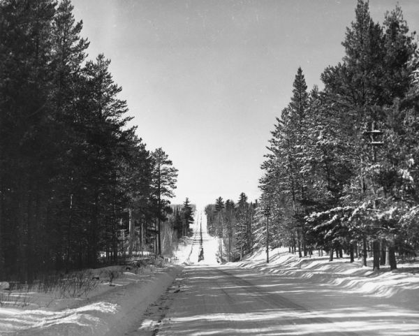 A snow-covered road disappears over a hill on a county highway between the Brule River and Lake Nebagamon. Pine trees line both sides of the road.