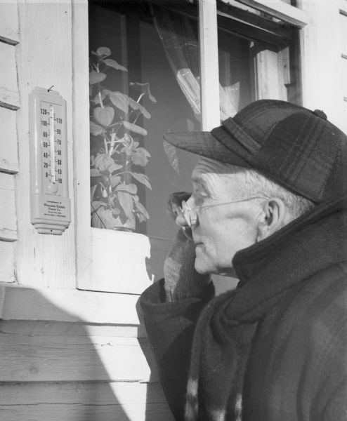 A man is peering over his glasses at a thermometer fastened to the outside of his house. He is dressed warmly in a plaid coat, scarf and hat. A houseplant is in the window behind him.