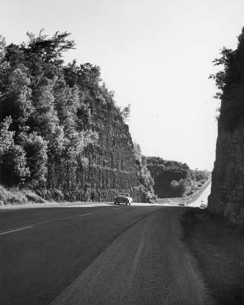 Two automobiles driving on a highway cutting between two halves of a rock outcropping. The rock was blasted to build the road.