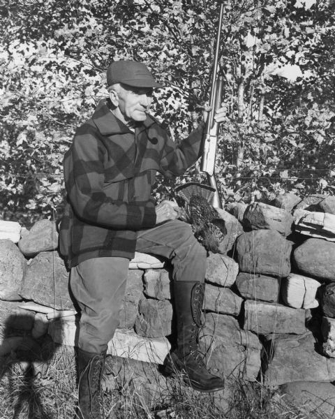 A hunter posing on a stone wall with his shotgun and the grouse he bagged. He is wearing a billed cap, plaid coat, tough trousers and lace-up knee-high boots. He is also smoking a pipe. In the background are trees. The shadow of the photographer's tripod and camera is in the lower left.