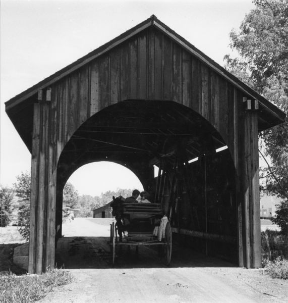 Rear view of a couple sitting in a horse-drawn buggy which is driving through a covered bridge at Stonefield Village. Buildings and trees are in the distance.