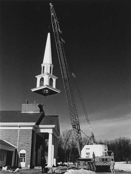 A crane setting a new steeple on the Pilgrim Congregational Church. People are standing near the crane on the ground, and men are standing on the roof helping the guide the crane.