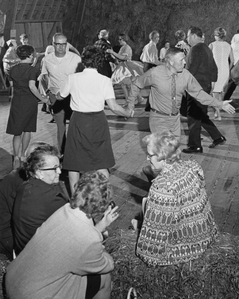 A crowd of various ages square dancing at a Barn Dance, Three women in the foreground are sitting on hay bales and talking.