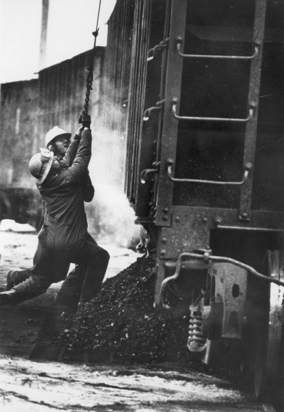 Two railroad workers are pulling on a chain next to a railroad car carrying coal.