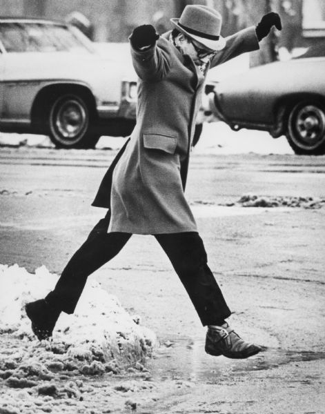 A warmly dressed man is leaping off a curb and over a puddle of slush while crossing the street. There are automobiles in the background.