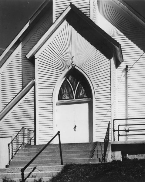 The entrance to a church with stained glass in the arch above the doors. The clapboard siding forms an interesting pattern in the peaks of the roof. Cement steps with iron railings lead up to the doors.