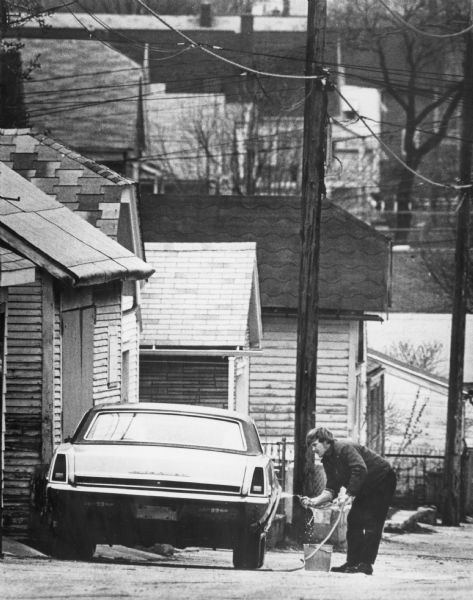 View down hill towards a man washing his car in the alley of a neighborhood. Rooftops are in the background.