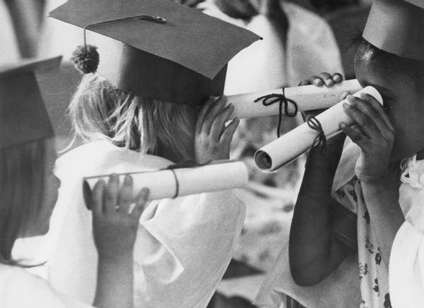 Grade school children are pretending their diplomas are binoculars in a graduation ceremony. They are wearing hand made robes, and caps with a pom pom instead of a tassel.