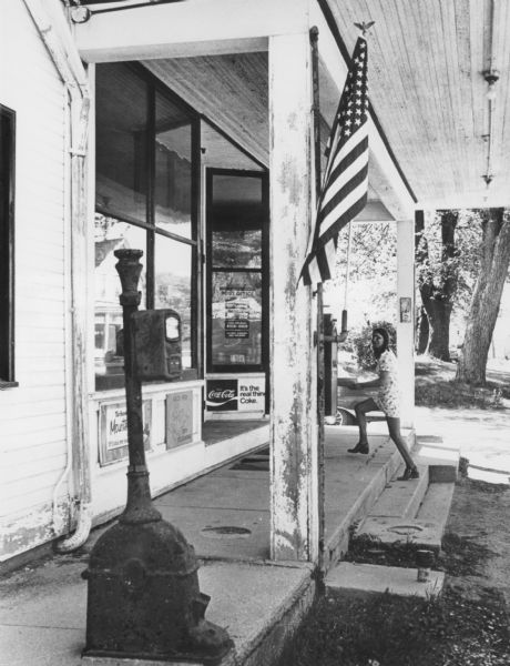 A teenage girl is walking up cement steps to a sidewalk in front of a country store and Post Office. An American flag is flying from a column in the foreground. Advertising signs are under the large front windows of the storefront. On the sidewalk in the left foreground is a box on a heavy-duty column supported by a large base, possibly a call box.