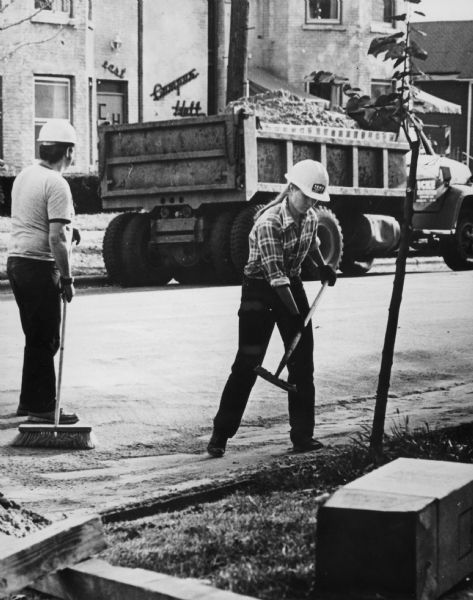 A woman and a man are working on a road construction crew, with a dump truck full of sand behind them. They are wearing work clothes and hard hats. In the background is a building with a sign that reads: "Campus Hall," with "east" over the door and the capital letters "C & H" on the door.  
