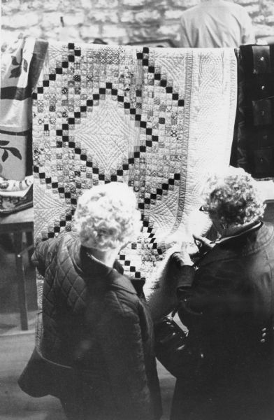 Two women are standing and admiring quilts hanging on a clothesline at a show or fair at Villa Louis.