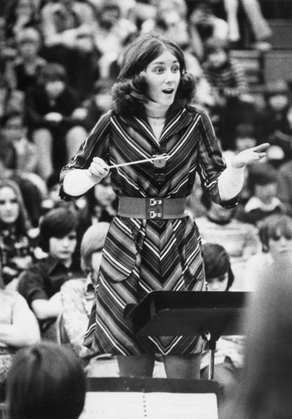 A young woman conducting a music group at a concert. Musicians are in the foreground, and in the background is an audience.