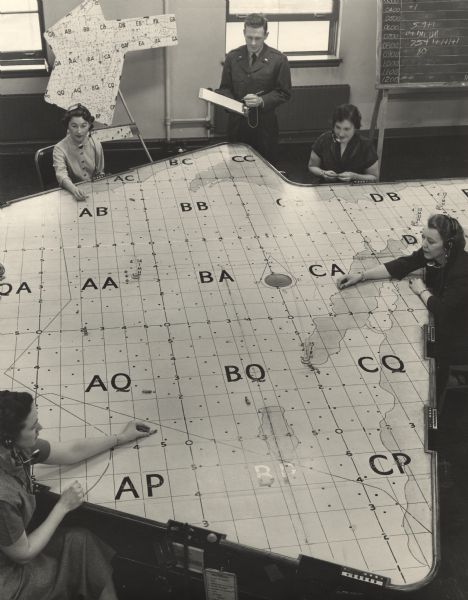 Elevated view of four women sitting, and one man standing, around a large map of northeast Wisconsin and Michigan's Upper Peninsula. The women are wearing headsets, and the man is wearing a uniform and is holding a clipboard.