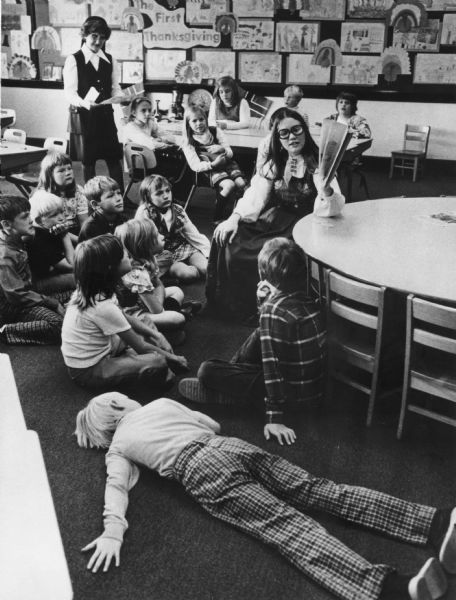 A group of children is seated on the floor and in chairs, listening to a teacher reading during story time. One boy is lying on the floor. A nun is standing in the upper left. The bulletin board on the wall displays student art about the First Thanksgiving.