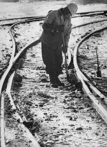 A railroad worker cleans snow out of a railroad track switch with a broom. He is dressed warmly with a hard hat.