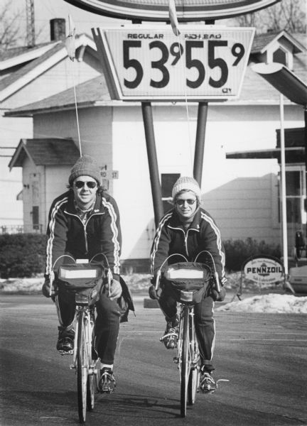 Two bicyclists on 10-speeds. They are wearing matching jackets and pants, with knit caps. The bikes have front bags on the handle bars. Note the price of gasoline on the sign above.