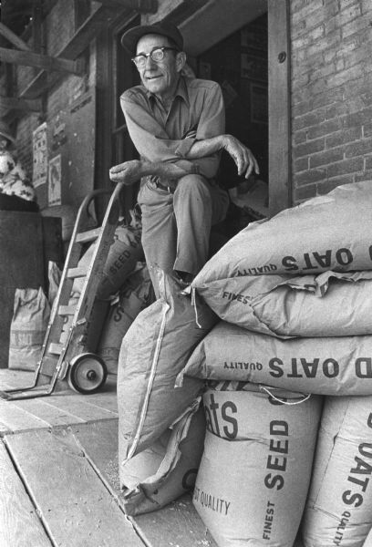 A man standing behind a pile of oats in bags on a loading dock. He is relaxing with one foot on the oats and his arms are crossed on his knee. Behind him are more bags of oats and salt with a hand cart leaning against them.