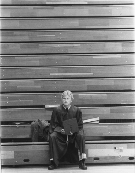 Male graduate wearing robes and sitting on the bottom row of empty bleachers holding his mortarboard.