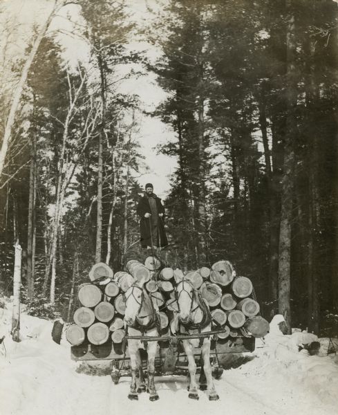 Ole Emerson's lumber property. A man is standing on top of a load of logs being pulled by a team of two horses on a sled through the snow. Caption from calendar: "Logging at Cable in 1906. A Bayfield county scene taken during the great years of the northern Wisconsin 'pineries'."