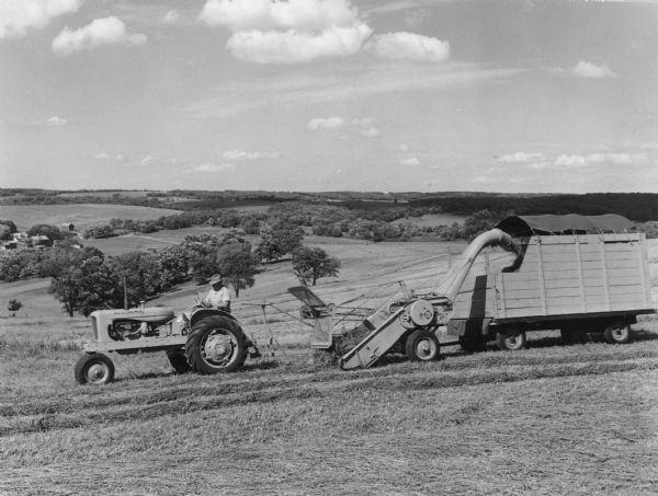 A man driving a tractor, pulling a chopper filling a forage wagon, over gently rolling hills.