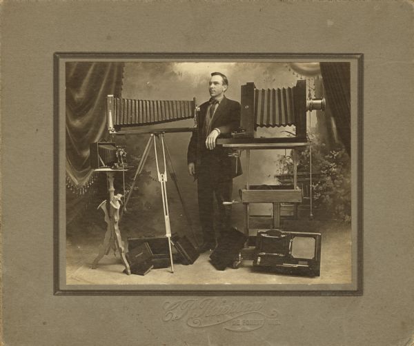 Full-length portrait of a man posed standing in front of a painted backdrop with a number of large format cameras and other photography equipment.