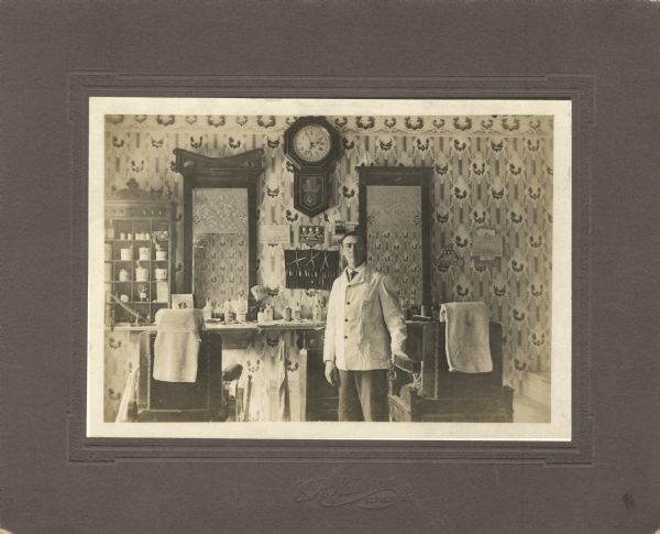 Man posed standing in a barber shop. His barbershop tools and equipment are behind him on a shelf under the two mirrors, in a cabinet on the left, and on the wall under advertisements. He is resting his hand on one of the barber chairs.