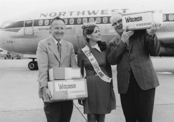 Group portrait of a woman standing between two men. In the background is a Northwest Airlines airplane. The woman is wearing a crown, and a sash that reads: "Alice in Dairyland." The men hold boxes labeled "Wisconsin Cheese."