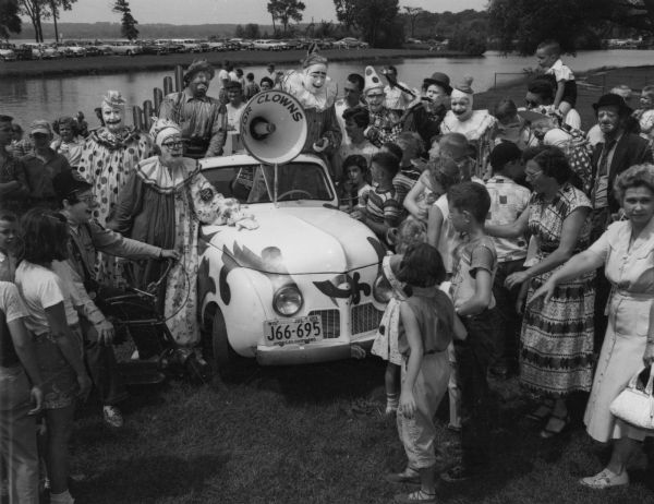 Group portrait of clowns standing beside and on a car decorated with the Zor Shriners logo. On the front of the car a large horn labeled "Zor Shrine" is mounted in the center of the windshield. On the left, one of the clowns is sitting on a bicycle or scooter. The clowns are surrounded by a large group of women and children. In the background is water, and on the opposite shoreline cars are parked near trees.