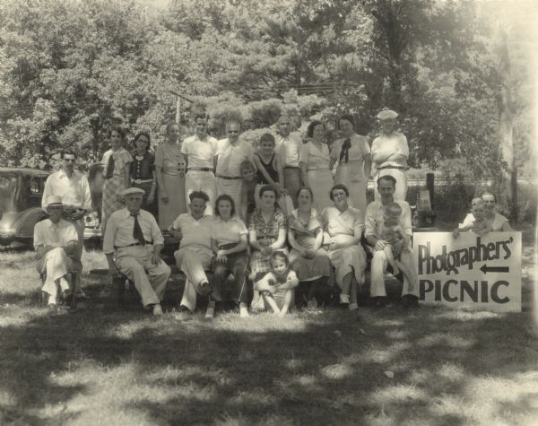 Group portrait of men, women and children posing outdoors under the shade of trees next to a sign that reads "Photographers' Picnic."