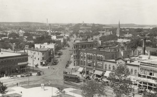 Elevated view looking down State Street taken from the Wisconsin State Capitol. On the right on Mifflin Street men are working on scaffolding on the facade of a building. A trolly is in the foreground on , and more are further down State Street. There is a horse-drawn carriage on Carroll Street. On the right is the Holy Redeemer Catholic Church located on West Johnson Street. There are smokestacks in the distance, and Bascom Hall and Lake Mendota are in the far background.