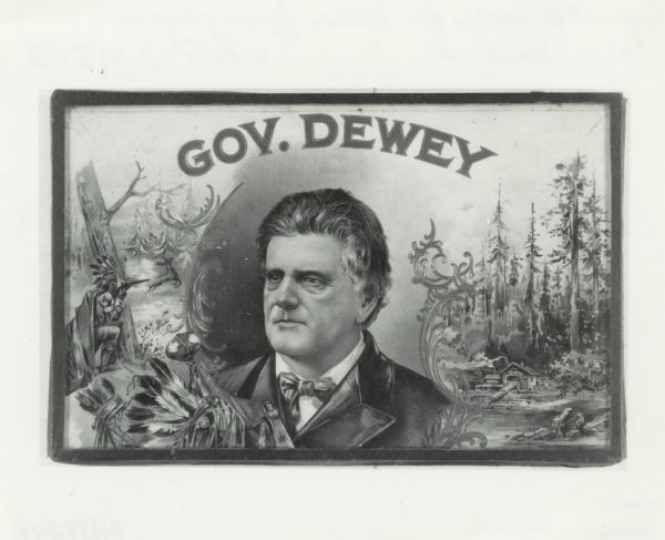 Cigar box cover with a head and shoulders portrait of Nelson Dewey. Beneath his portrait is a Native American headdress, a tomahawk, a stone club and a quiver of arrows. In the background on the right is a depiction of a cabin in a forest on a shoreline. On the left is a Native American man behind a tree trunk pointing a rifle at a running deer.