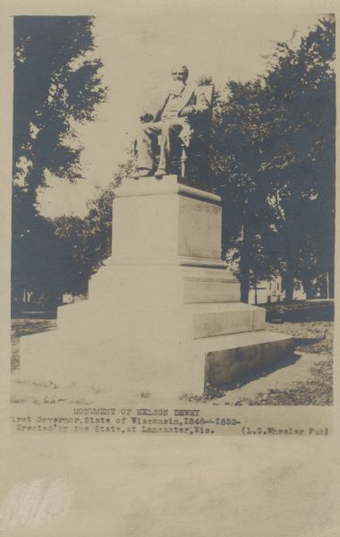 Postcard depicting the monument of Nelson Dewey. He has a long beard and sits in a chair on a pedestal. Trees are  in the background. Text at the bottom reads: "Monument of Nelson Dewey. First Governor, State of Wisconsin, 1848--1852-. Erected by the State, at Lancaster, Wis."