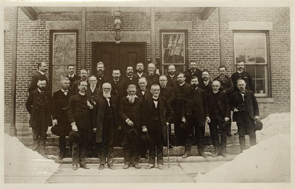 Group portrait of men standing under the porch of a brick building. Nelson Dewey is the second man from the right in the front row with a long white beard and holding a hat.