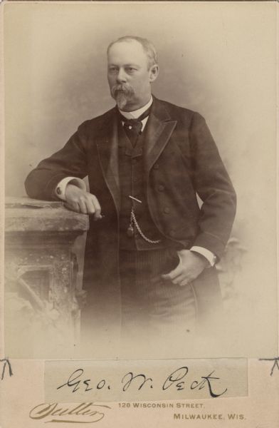 Three-quarter length portrait in front of a painted backdrop of George Peck on a cabinet card. He is wearing a suit and necktie and is resting his right forearm on a stone pillar. A watch fob is attached to his vest.