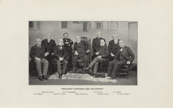 President Harrison sits in the middle of the group  on an ornate chair on a slight dais covered with a rug. His cabinet members sit on his left and right. Behind the group of men are portraits of men hanging on the wall. From left to right are: S.B. Elkins; John W. Noble; John W. foster; Jno. Wanamaker; Benjamin Harrison; B.F. Tracy; Charles Foster; Jeremiah M. Rusk; W.H.H Miller.