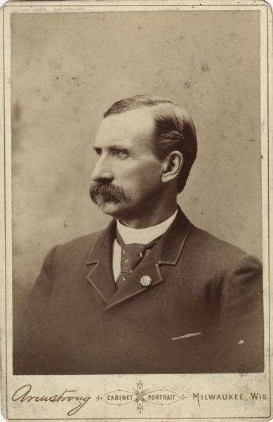 Semi-profile portrait on a cabinet card of William Dempster Hoard, looking to the left. He is wearing a suit, a necktie with small angled rectangles, and a pin that bears a resemblance to the American flag.