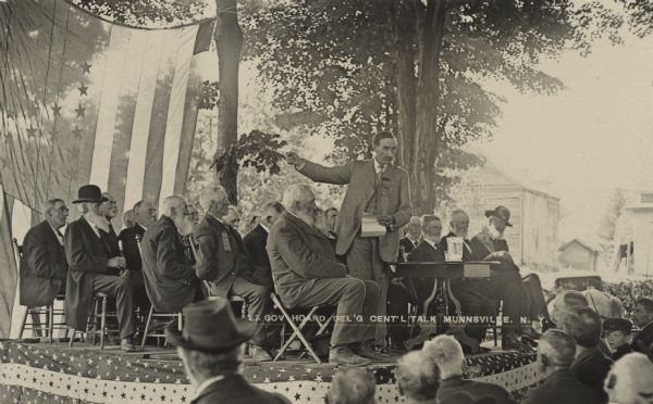 Ex-governor William Dempster Hoard standing on a stage in front of an audience. He holds a piece of paper in his left hand, and is gesturing with his right arm. Behind him on the stage are a group of men sitting in chairs. A large American Flag is tied to trees behind the stage. Caption reads: "Ex. Gov. Hoard Del'g Gen'l Talk, Munnsville, N. Y."
