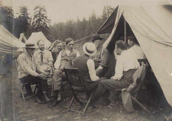 A group of men sit in chairs close together around the entrance of an open tent. In the background are trees and more tents. The men are wearing shirts, boots, vests and brimmed hats (one man wears a tie). George Peck sits facing towards the camera (third in from the right, in between two men with their backs to the camera).