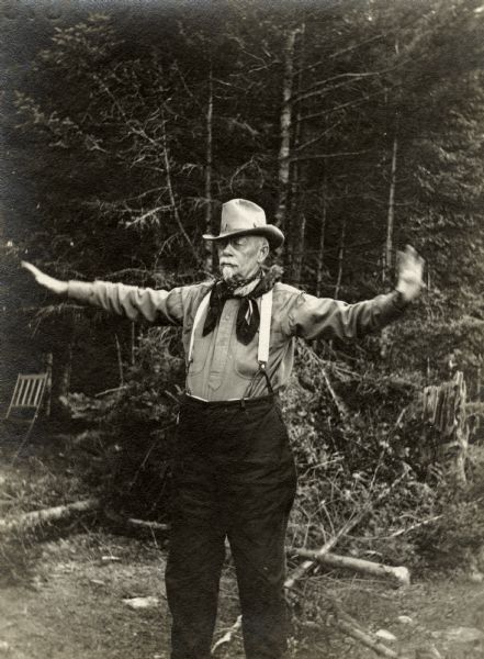 Former governor George W. Peck stands in the woods with his arms outstretched. He is wearing a brimmed hat, shirt, neck scarf, suspenders, and pants.