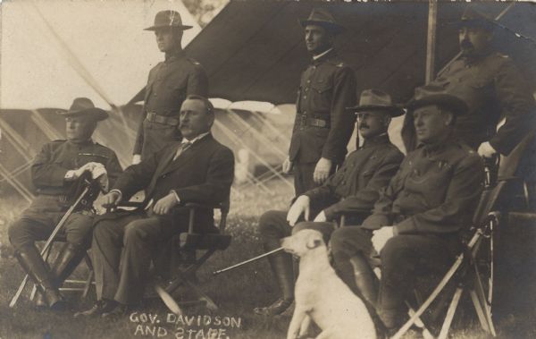 Governor James O. Davidson (sitting second from the left), outdoors in front of a tent with six other men, three standing and three sitting. There is a white dog sitting in the foreground. Davidson wears a suit and tie. The other men are dressed in military uniforms, tall boots, brimmed hats, white gloves, with their swords by their side.